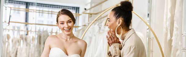 Een jonge brunette bruidsmeisje en een mooie bruid in een witte jurk naast elkaar staan voor een spiegel. - Foto, afbeelding