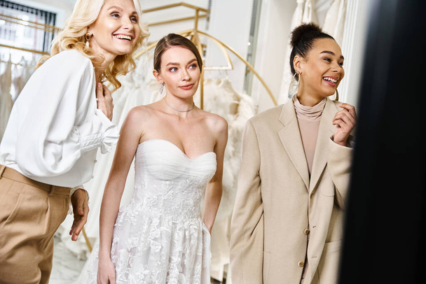 Tři ženy, mladá brunetka a ohromující nevěsta v bílých šatech stojí vedle sebe před zrcadlem. - Fotografie, Obrázek
