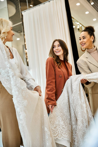 Une jeune mariée rayonnante, sa mère et sa meilleure amie debout ensemble, faisant du shopping pour son mariage. - Photo, image