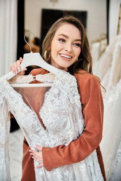 Una giovane donna regge un vestito in un negozio immerso nella gioia dello shopping di nozze. - Foto, immagini