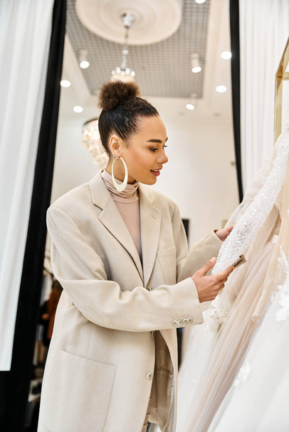 Uma jovem noiva bonita de pé ao lado de um vestido de noiva branco, em uma boutique nupcial. - Foto, Imagem