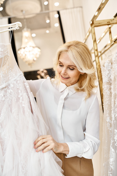 Красивая помощница продавца средних лет просматривает свадебные платья на стойке в свадебном салоне. - Фото, изображение