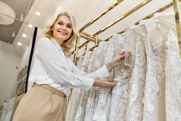Egy középkorú, gyönyörű bevásárló asszisztens áll az esküvői szalonban egy ruhafogas előtt, segítve az ügyfeleket.. - Fotó, kép