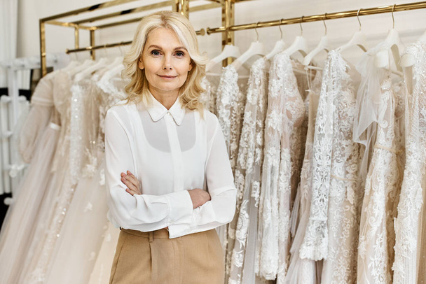 Un hermoso asistente de compras de mediana edad se para ante un estante de elegantes vestidos de novia en un salón de novias.. - Foto, imagen