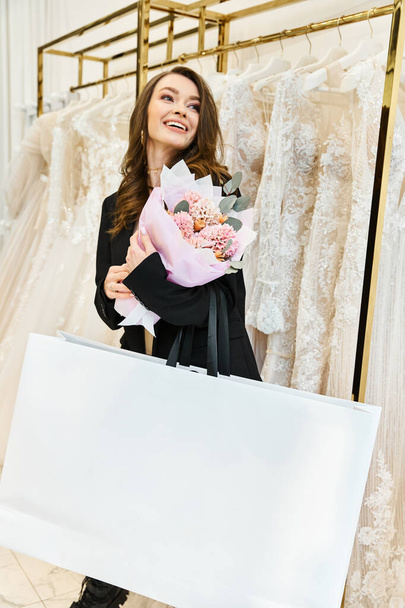 Jovem noiva morena segurando buquê na frente do rack de vestidos de noiva no salão de casamento. - Foto, Imagem