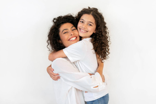 Happy young mother hugging her daughter and smiling at camera, expressing tenderness, love and care, standing over white studio wall background - Photo, Image