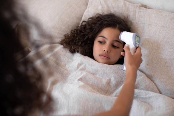 Cuidar a la madre midiendo la temperatura de su hija usando termómetro infrarrojo sin contacto, niña acostada en la cama debajo de la manta - Foto, imagen