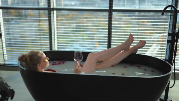 Relaks w spa. Kobieta leży w wannie z kieliszkiem z czerwonym winem. Dziewczyna relaksuje się w łazience w domu. Ładna kobieta pijąca wino w gorącej kąpieli. Zmniejszenie stresu. Odpoczynek po ciężkim dniu pracy - Materiał filmowy, wideo