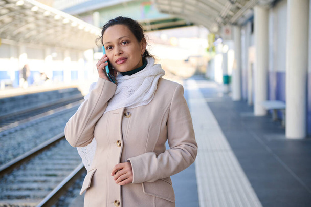 Mujer joven multiétnica hermosa en sus 30 años está utilizando un teléfono móvil, conversando mientras espera un tren en una plataforma de la estación de tren. Personas, tecnologías y transporte urbano público - Foto, Imagen