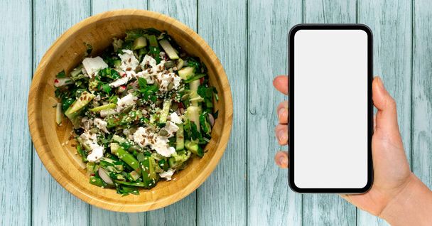 Nutrition mobile App-Attrappe. Smartphone mit leerem Bildschirm in der Hand neben Holzschüssel mit grünem Salat. - Foto, Bild