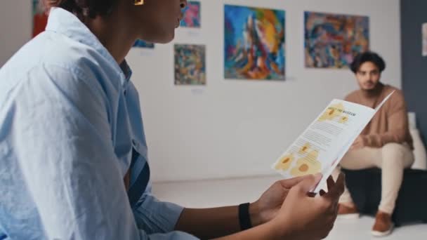 Medium close-up kantelen schot van handen en gezicht van jonge krullende Afro-Amerikaanse vrouw zittend op de bank in de kunstgalerij op moderne tentoonstelling en het lezen van informatie folder - Video