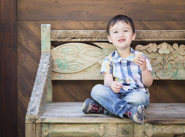 Симпатичный мальчик смешанных рас сидит на скамейке и ест сэндвич
 - Фото, изображение