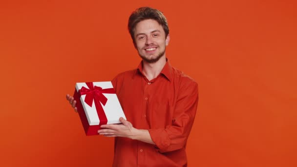 Pozytywny uśmiechnięty biały mężczyzna prezentujący pudełko prezentów urodzinowych rozciąga się ręce oferują zapakowany obecny bonus kariery świętując promocję partii wyprzedaży rabatowej. Przystojniak odizolowany na pomarańczowym tle - Materiał filmowy, wideo