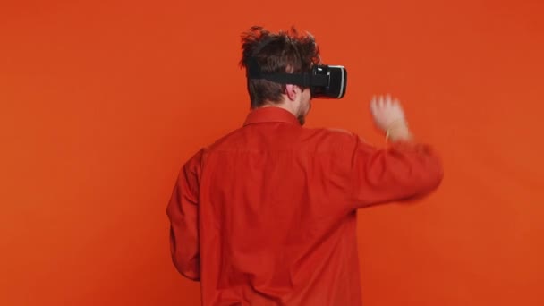 Tylny widok z tyłu podekscytowany szczęśliwy człowiek za pomocą aplikacji kasku słuchawkowego do gry symulacyjnej. Oglądanie wirtualnej rzeczywistości wideo 3D 360. Młody facet w okularach VR odizolowany na pomarańczowym tle. Technologia przyszłości - Materiał filmowy, wideo