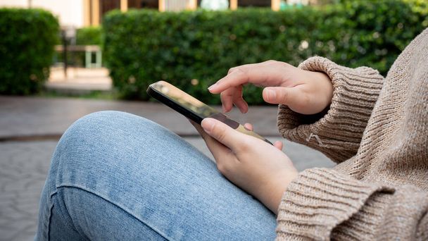 Foto ritagliata di una donna in un accogliente maglione seduta su una panchina in un giardino del parco cittadino e utilizzando il suo smartphone. persone e concetti di stile di vita - Foto, immagini