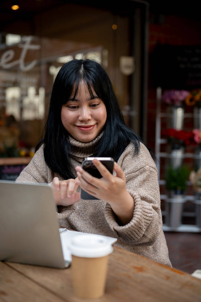 A positive, beautiful Asian woman in a cosy sweater is responding to messages on her smartphone while working remotely at a coffee shop in the city. People, wireless technology, and city life concepts - Photo, Image