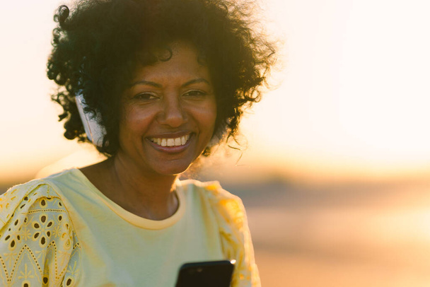 Mulher africana sorrindo enquanto ouve música com celular ao ar livre durante o pôr do sol - Foto, Imagem