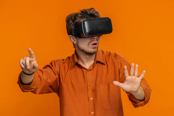 Возбужденный бородатый человек использует гарнитуру шлем приложение для игры моделирования. Просмотр виртуальной реальности 3D 360 видео воображения. Молодой парень в защитных очках изолирован на оранжевом фоне в помещении. Технологии будущего - Фото, изображение