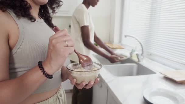Schiefe Aufnahme einer jungen Frau, die Müsli zum Frühstück isst, während ihr Freund im Hintergrund Geschirr spült - Filmmaterial, Video