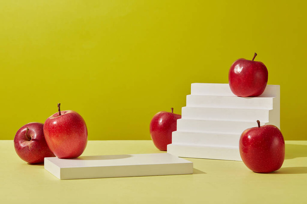 Προϊόν και προώθηση έννοια για τη διαφήμιση καλλυντικό προϊόν του εκχυλίσματος μήλου. Μερικά ώριμα κόκκινα μήλα διακοσμημένα σε πράσινο φόντο με ορθογώνιο λευκό βάθρο και λευκή σκάλα. Πρόσθια όψη - Φωτογραφία, εικόνα