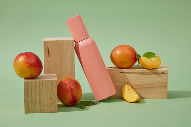 Mockup scene voor reclame en branding cosmetische van perzik (prunus persica) extract. Een roze cosmetische fles versierd met verse perziken en houten blokken op een groene achtergrond. Ruimte voor ontwerp - Foto, afbeelding