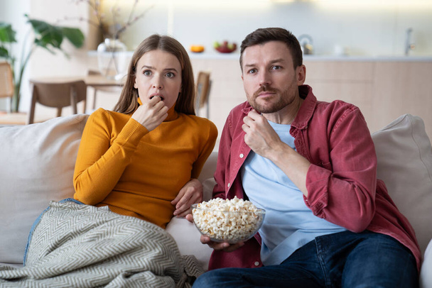 Zaskoczona rodzinna para ogląda telewizję, film, serial siedzący na kanapie w domu jedząc popcorn. Niesamowity zrelaksowany żonaty mężczyzna i kobieta cieszyć się rozrywką razem zainteresowany thrillerem z ekscytującej fabuły - Zdjęcie, obraz