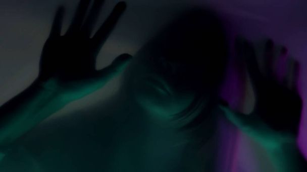 Blurred silhouette of a woman behind a mat curtain, in pink and green neon light, close up. A woman is scraping her fingernails on the barrier, trying to tear it. The concept of ghosts and spirits - Foto, afbeelding