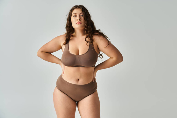 attractive plus size young woman in underwear with brown hair posing with hands on grey background - Valokuva, kuva