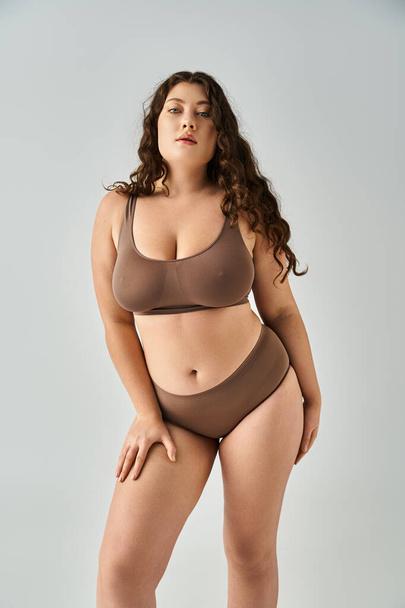 beautiful curvy woman in underwear with brown hair leaning forward and posing on grey background - Foto, Bild