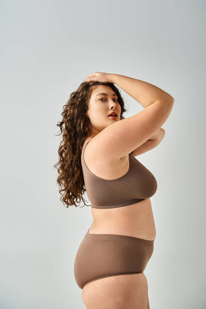 attractive curvy woman in underwear with curly brown hair putting hands on head on grey background - 写真・画像
