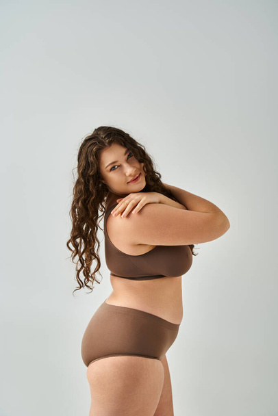 beautiful plus size woman in underwear with curly brown hair hug herself sideways on grey background - Photo, Image