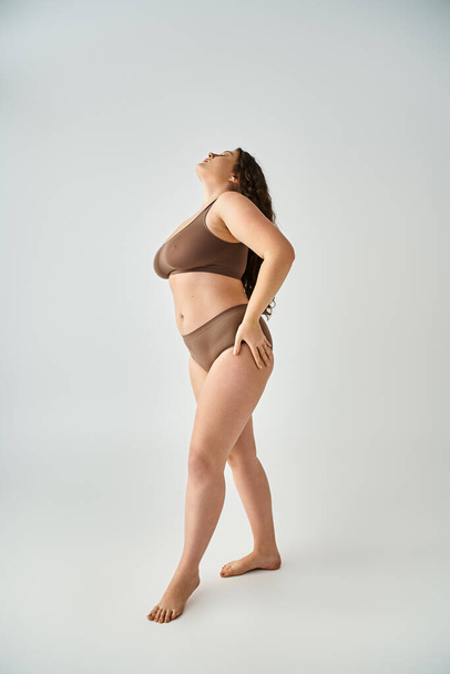 aantrekkelijke plus size vrouw in ondergoed met handen op heupen zetten hoofd naar achteren in grijze achtergrond - Foto, afbeelding