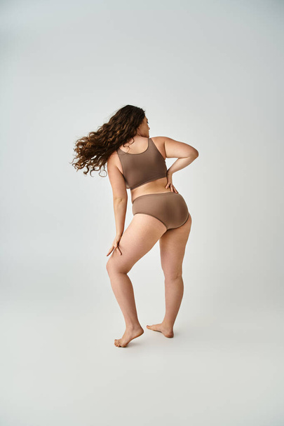 plus size young woman in underwear with curly brown hair posing from back on grey background - Фото, изображение