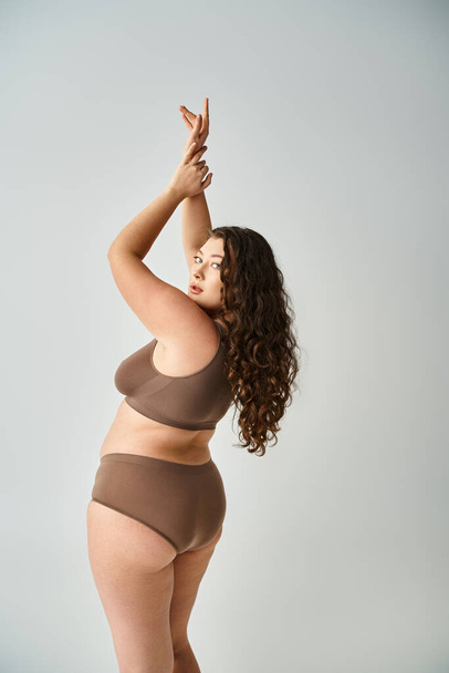 attractive curvy woman in underwear with curly brown hair posing from back and putting hands to up - Foto, Bild