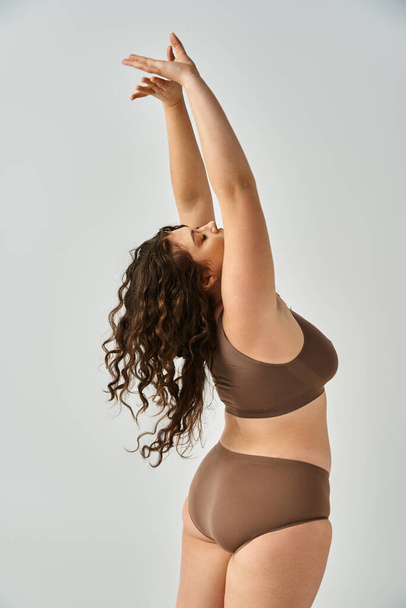 charming curvy young woman in underwear with curly brown hair from back putting hands to up - Foto, Imagem