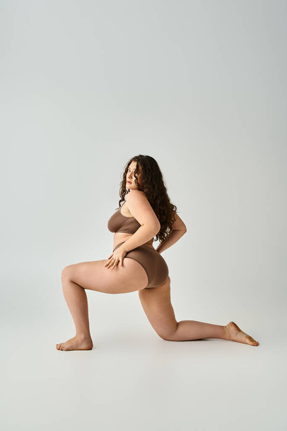attractive plus size woman with curly brown hair posing from sideways with legs on grey background - Foto, imagen