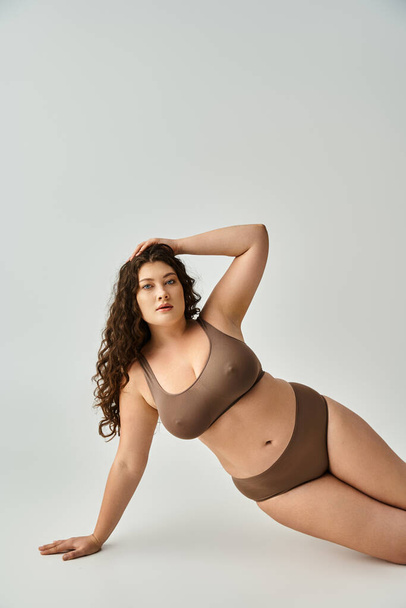 alluring plus size woman in lingerie with curly brown hair leaning on arm and putting hand on head - 写真・画像