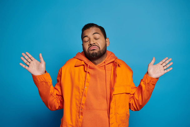 charismatic african american man in orange outfit showing that knows nothing on blue background - Photo, Image