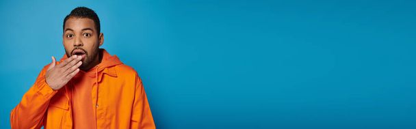 pancarta del hombre afroamericano sorprendido en traje naranja que cubre la boca con la mano sobre fondo azul - Foto, imagen