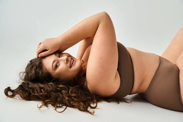 glimlachende curvy jonge vrouw in bruine lingerie met krullend haar liggen en het zetten van de hand op hoofd - Foto, afbeelding