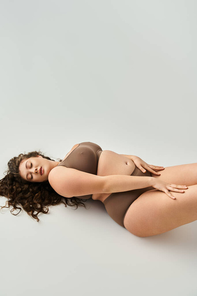 alluring plus size young woman in brown underwear with hands on hips and closed eyes - Фото, зображення