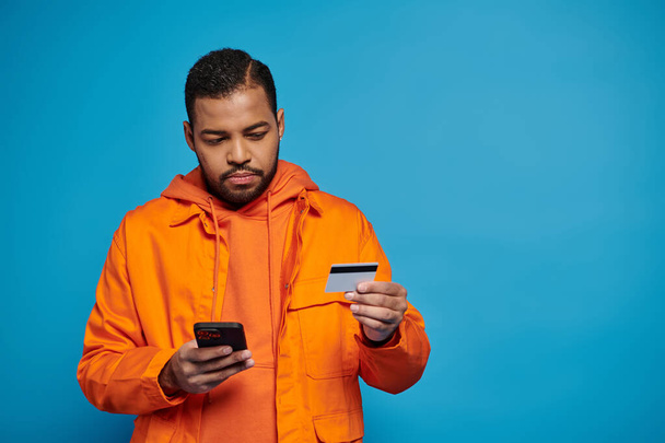 ориентированный африканский американский молодой парень в оранжевом костюме платит онлайн покупки на синем фоне - Фото, изображение
