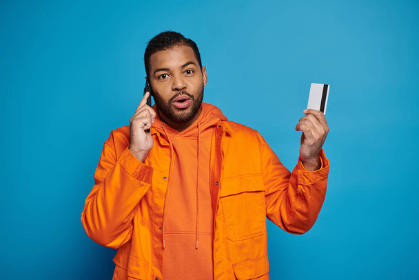 sorprendido afroamericano joven llamando en smartphone con tarjeta de crédito sobre fondo azul - Foto, imagen