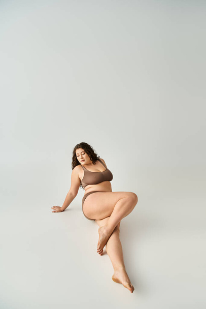 svůdná plus velikost mladá žena v hnědém spodním prádle sedí, naklání se k pažím a dává nohu - Fotografie, Obrázek