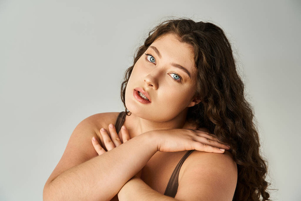 portrait of curvy woman in her 20s with curly brown hair and blue eyes covering hands to neckline - Фото, зображення