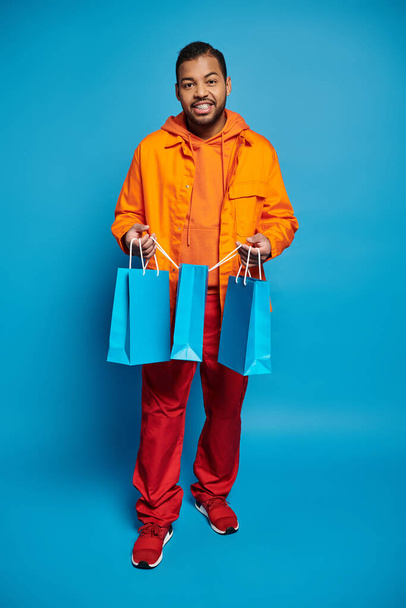 cheerful african american man in orange outfit holding shopping bags against blue background - Photo, image