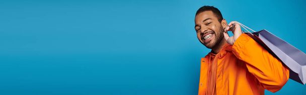 banner of african american man in orange outfit with shopping bag over shoulder on blue background - Photo, Image