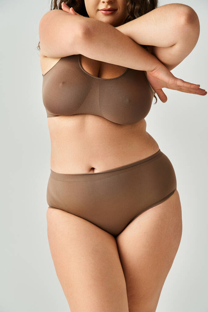 cropped shot of body plus size young woman in brown lingerie crossing arms to up on grey background - Photo, Image