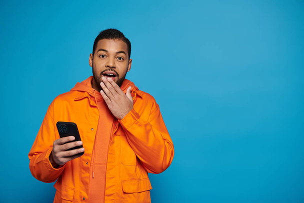 sorprendido hombre afroamericano en traje naranja desplazándose en las redes sociales y cubriendo mano a boca - Foto, imagen
