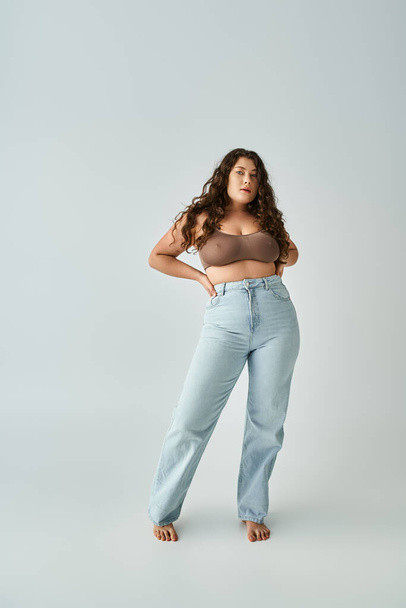lovely curvy girl in brown bra and blue jeans posing with hands in pockets on grey background - Photo, Image